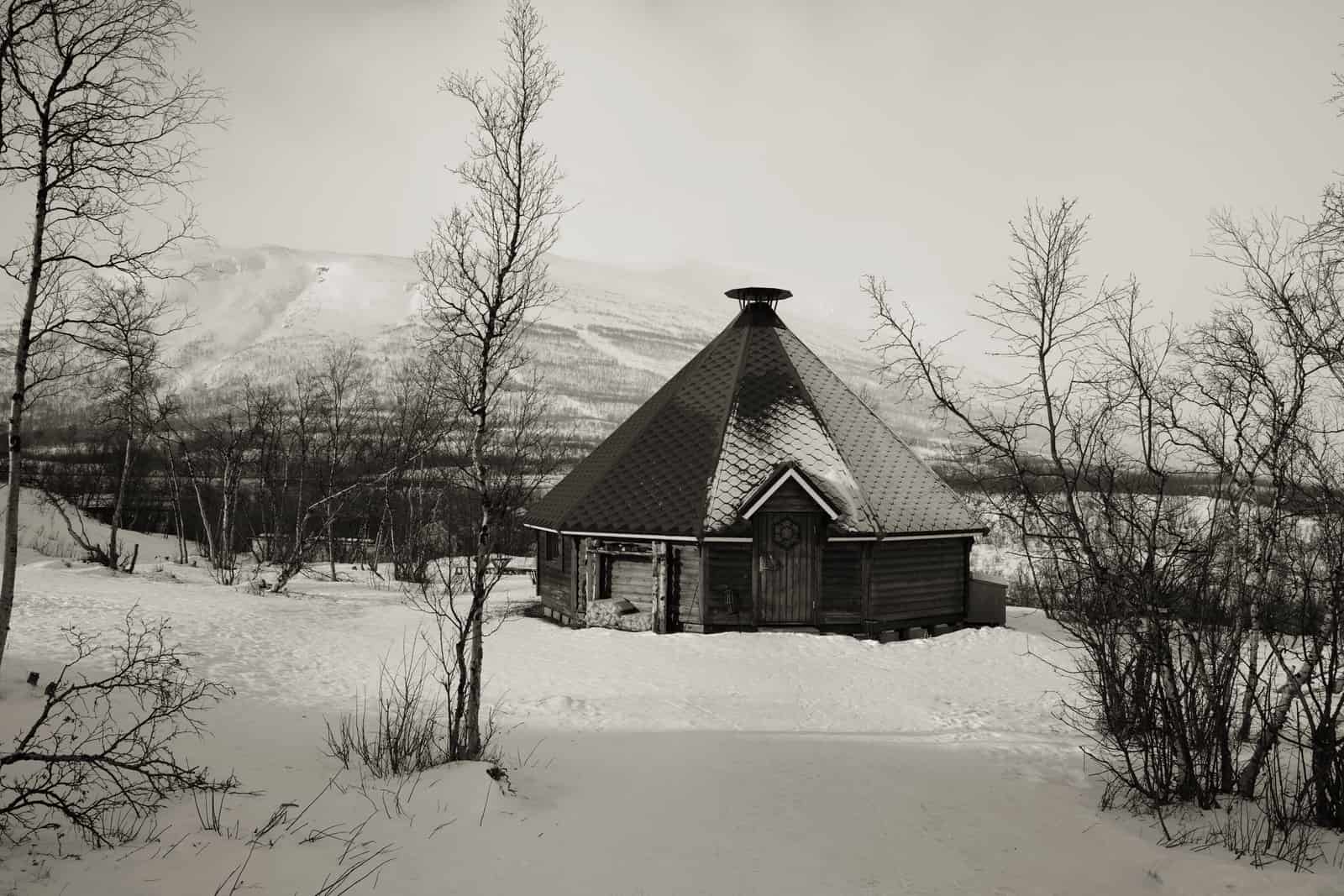A wooden sauna cabin standing among snow-covered trees in low winter light